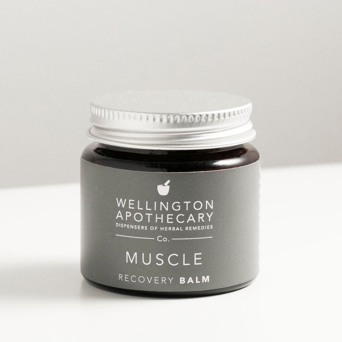 Amber glass jar with aluminium lid on white bench, labeled as muscle recovery balm. 