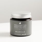 Amber glass jar with aluminium lid on white bench, labeled as muscle recovery balm. 
