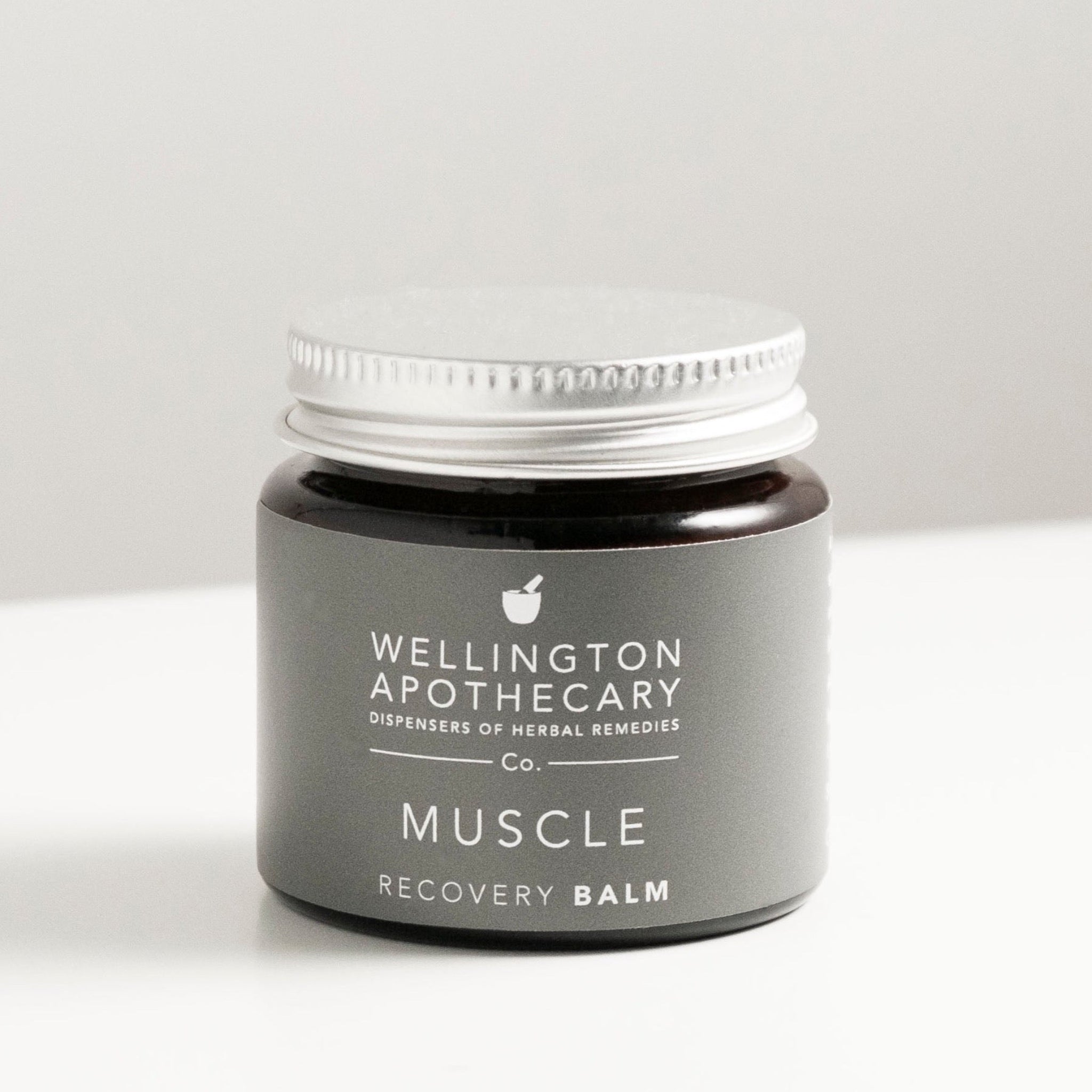 Amber glass jar with aluminium lid on white bench, labeled as muscle recovery balm. 