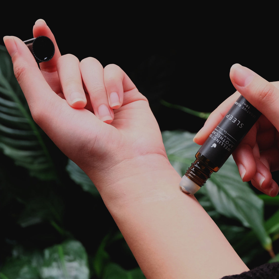 An amber glass bottle of Sleep aromatherapy blend being rolled onto a wrist with dark green leaves in the background. 